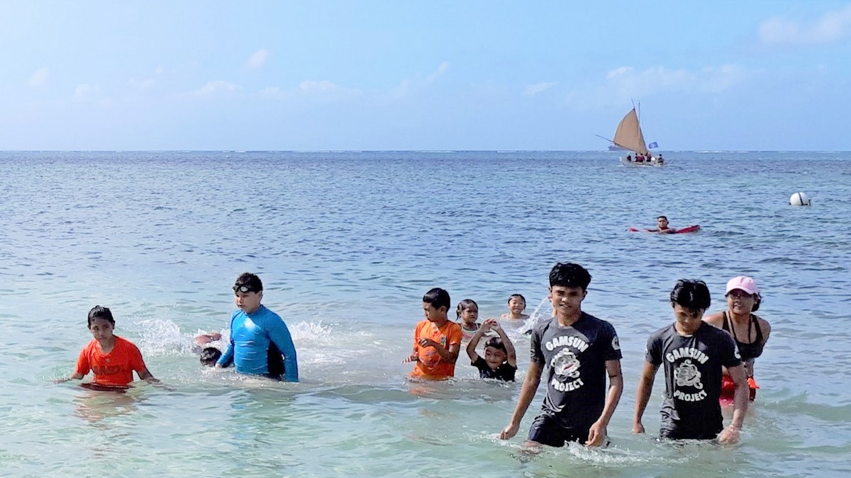 Gamsun Project youngest class participants, foreground, are seen with lifeguards while PSS Co-Op participants sail Anåguan, background. Photo by Andrea Carr